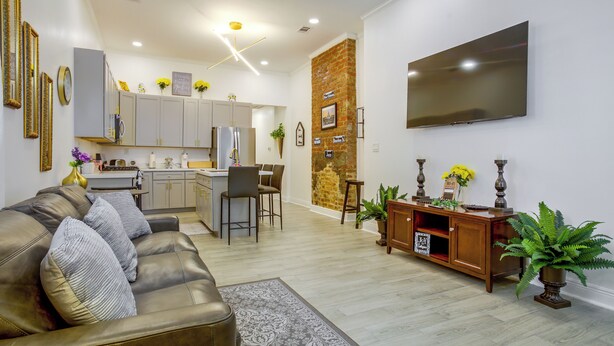 Una sala de estar moderna con un sofá de cuero, una zona de comedor con taburetes de bar, una cocina con armarios grises y una pared de ladrillo decorativa.