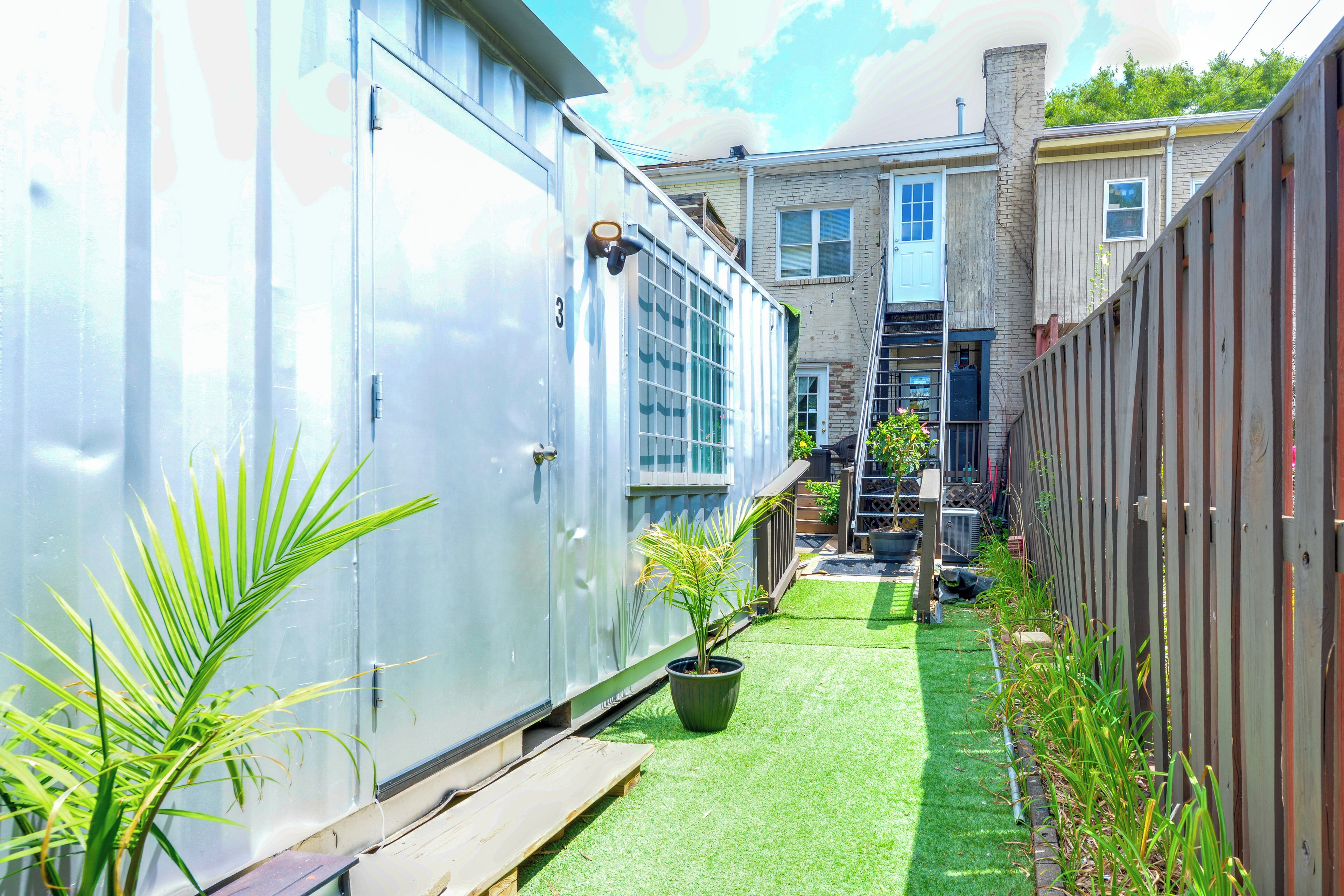 A narrow alleyway with a green artificial turf path, metal storage units, and a wooden fence on one side.