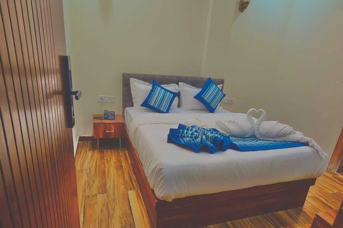 A bedroom with a wooden floor, a bed with white bedding and blue pillows, a bedside table, and a wall-mounted light fixture.