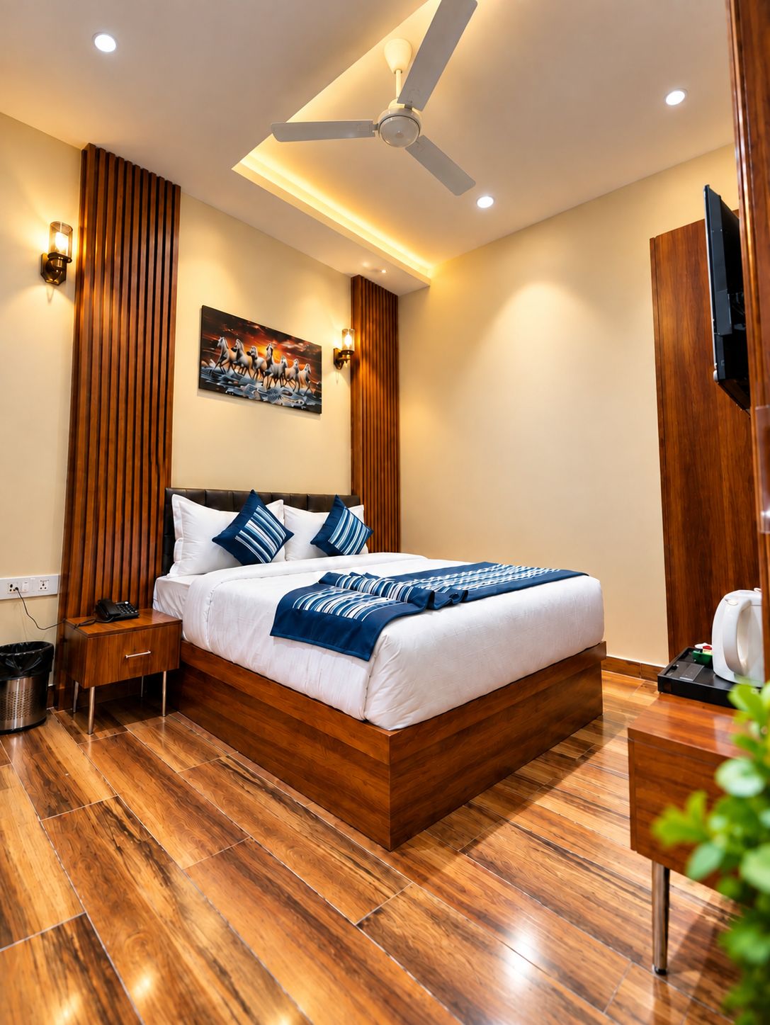 Una habitación de hotel con piso de madera, una cama con ropa de cama blanca y detalles azules, un ventilador de techo y un cuadro en la pared.