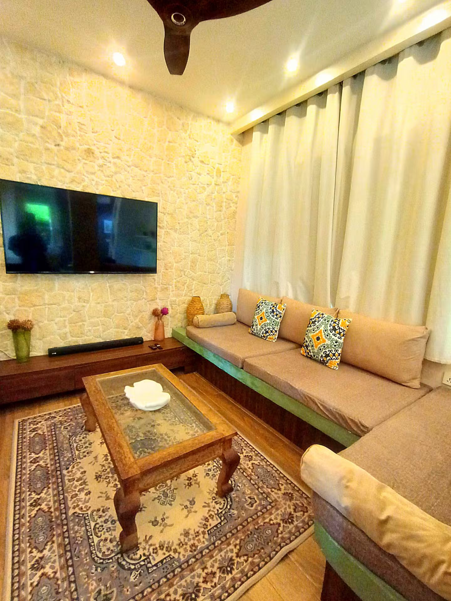 Ein Wohnzimmer mit einer Steinsäule, einem Flachbildfernseher, einem Sofa mit gemusterten Kissen, einem Couchtisch und einem Teppich mit Muster.