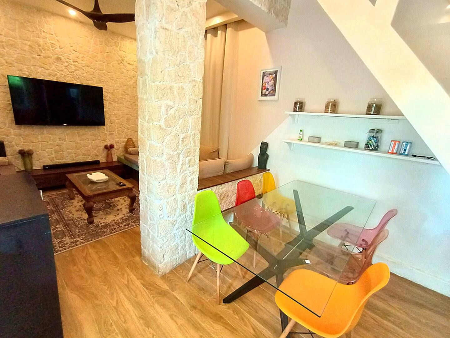 Un salon moderne avec un mur en pierre, un téléviseur à écran plat, une table à manger en verre entourée de chaises colorées et une table basse en bois.