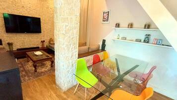 Un salon moderne avec un mur en pierre, un téléviseur à écran plat, une table à manger en verre entourée de chaises colorées et une table basse en bois.