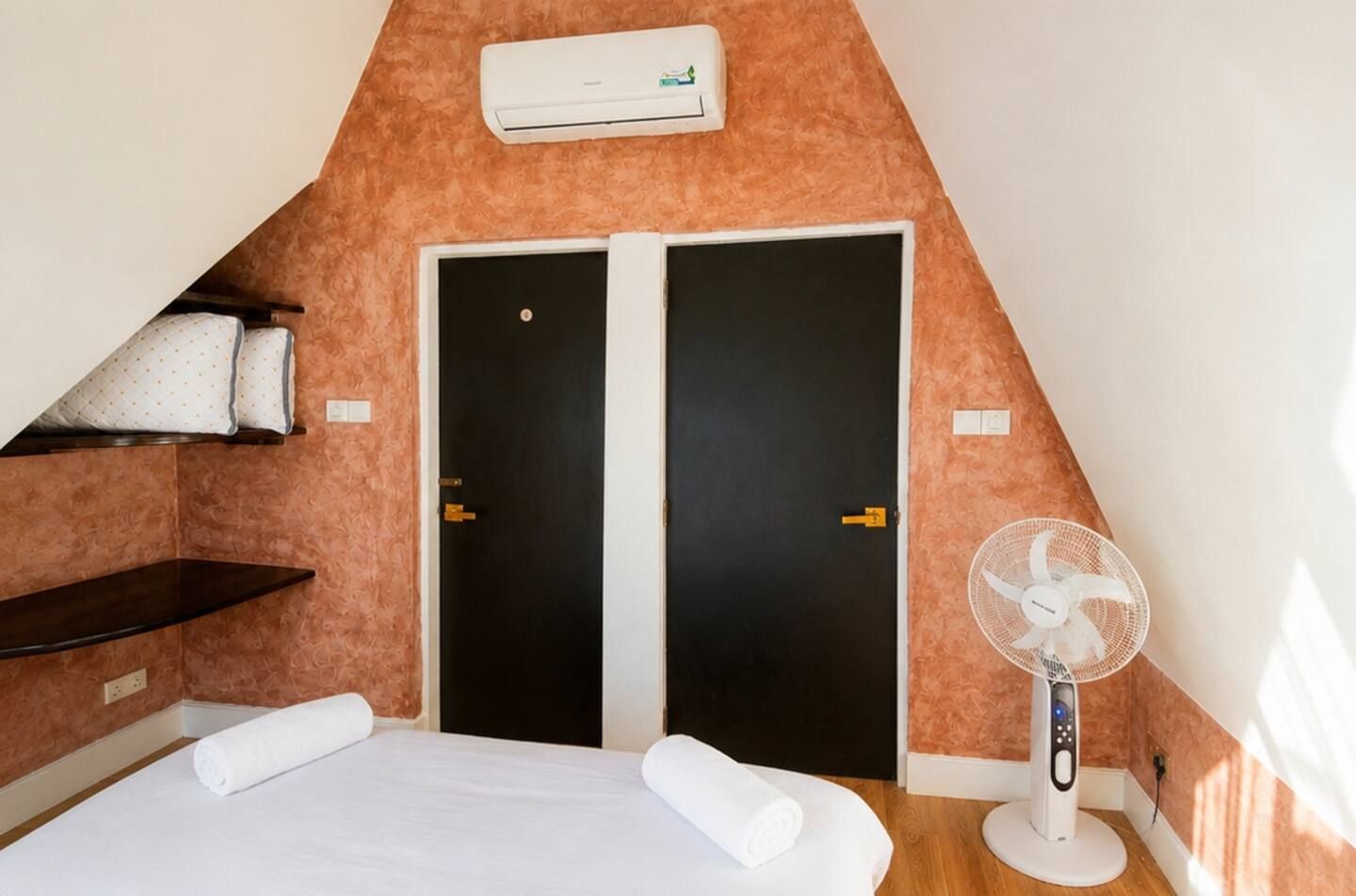 Une chambre avec un plafond en pente, deux portes noires, un lit blanc avec des serviettes roulées, une table de chevet, un climatiseur blanc et un ventilateur sur pied.