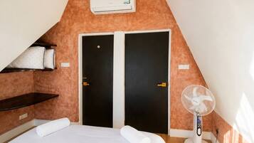 Une chambre avec un plafond en pente, deux portes noires, un lit blanc avec des serviettes roulées, une table de chevet, un climatiseur blanc et un ventilateur sur pied.