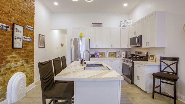 A modern kitchen with white cabinetry, a marble island, and a brick accent wall. - Stylish Unit 3 Mi to French Quarter & Audubon Park (New Orleans)
