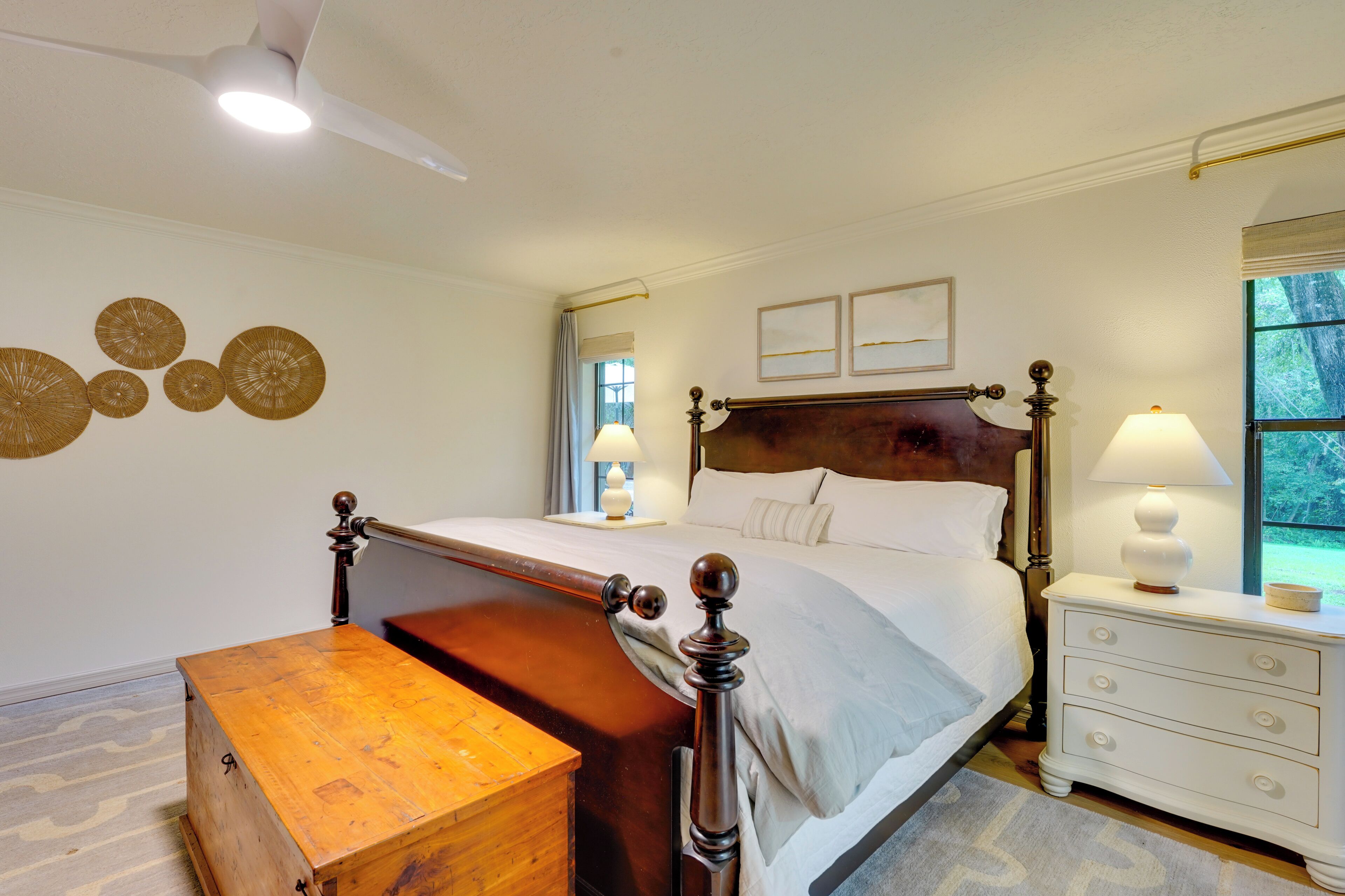 Un dormitorio con cama de madera, mesitas de noche, un aparador, un baúl de madera, un ventilador de techo y decoración en las paredes.