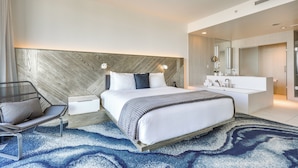 Een moderne hotelkamer met een groot bed, nachtkastje, stoel, ingebouwde kaptafel met spiegel en een blauw vloerkleed met patroon.