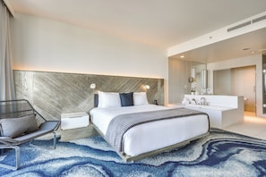 Ein modernes Hotelzimmer mit einem großen Bett, einem Nachttisch, einem Sessel, einer integrierten Schminkkommode mit Spiegel und einem blauen Teppich mit Muster.