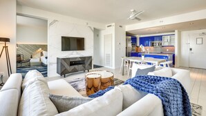 Een moderne woonkamer met een bank, salontafel en een open haard. Er is een keuken met blauwe kasten en een eettafel.