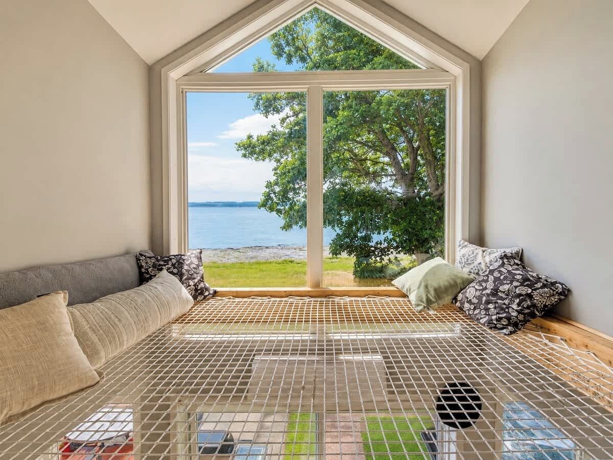 A room with a large window, a metal grid floor, a sofa with cushions, and a view of a body of water and trees.