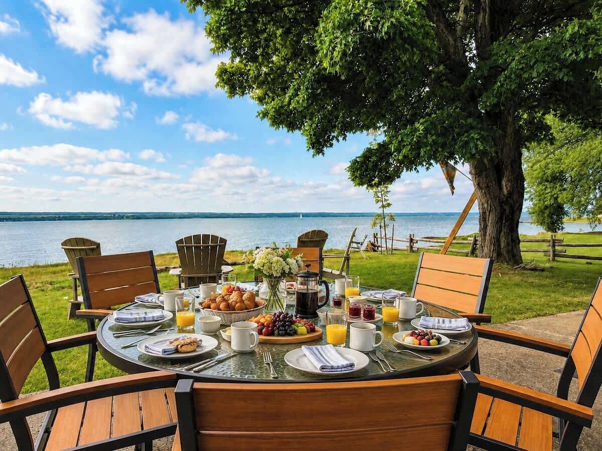 A round table set for breakfast with chairs, a view of the water, and a tree in the background.