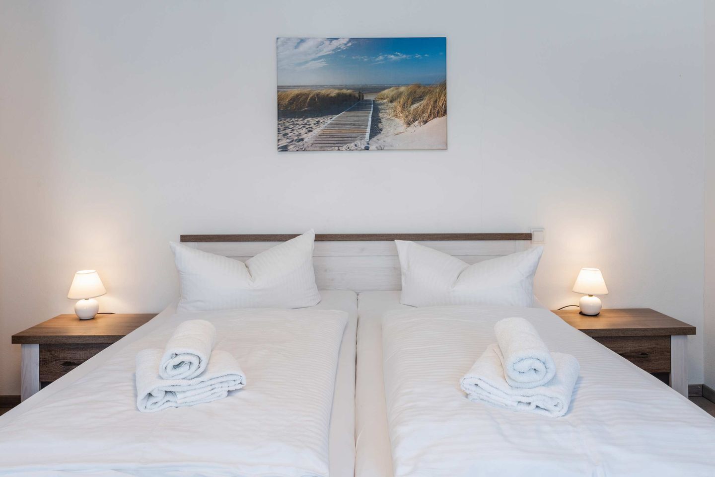 A double bed with white linens and pillows, two bedside tables with lamps, and a painting of a beach scene on the wall.