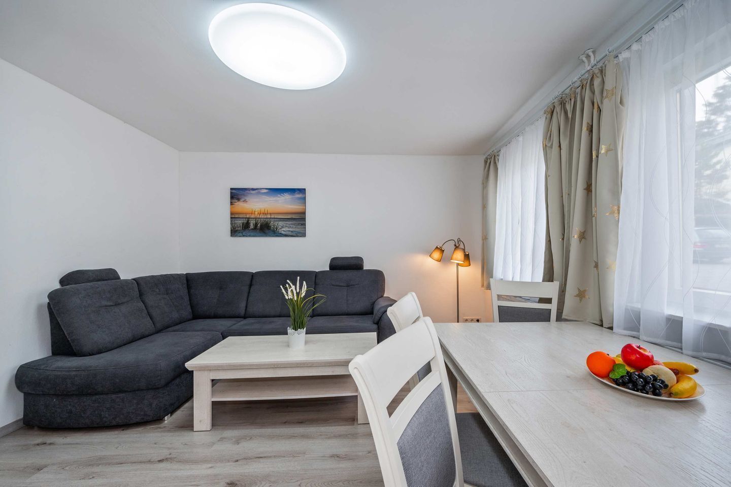 A modern living room with a sectional sofa, a dining table with chairs, a coffee table, a floor lamp, and a painting on the wall.