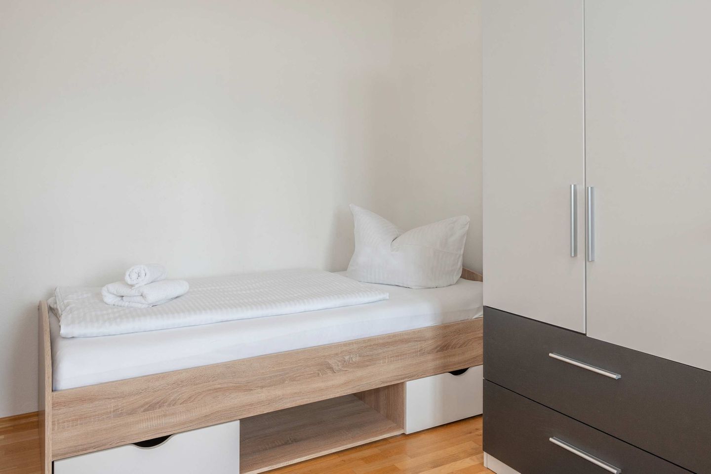 A single bed with a wooden headboard, white bedding, and a rolled towel on the side. There is a wardrobe with two drawers and a cabinet with a handle.