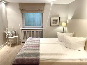 A hotel room with a bed, a chair, a radiator, a window with curtains, a lamp, and a framed picture on the wall. - Haus Wattblick - Haus Wattblick Apartment WB/01 (Sylt)