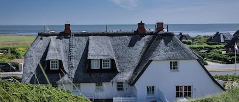 A traditional thatched-roof house with a white facade, situated on a grassy hill overlooking a coastal area.
