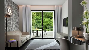 A modern hotel room with a sofa, a small table with a book, a lamp, a chair, and a view of greenery through a large sliding door.