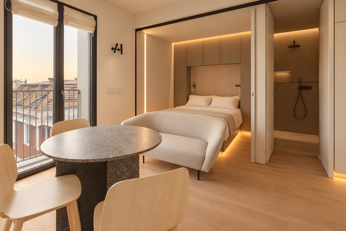 A modern hotel room with a bed, a round table, and chairs, a shower area, and a view of the cityscape.