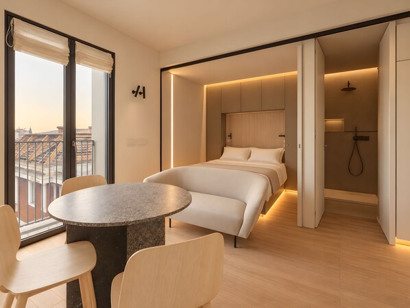 Quarto de hotel moderno com cama, mesa redonda e cadeiras, área de chuveiro e vista para a paisagem urbana.