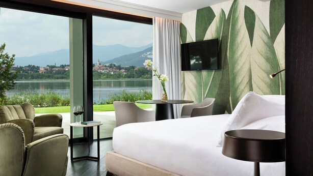 A hotel room with a bed, a TV mounted on the wall, a view of a lake and mountains, and a dining area with chairs and a table.