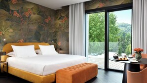 A modern bedroom with a large bed, a wooden bench, a dining table with a vase, and a balcony with a view of greenery.