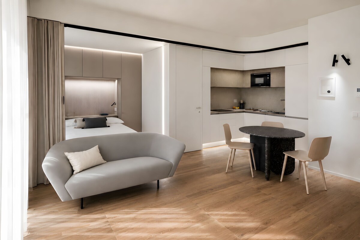 A modern living room with a sofa, a dining table with chairs, a kitchen area with a microwave, and a large window with curtains.
