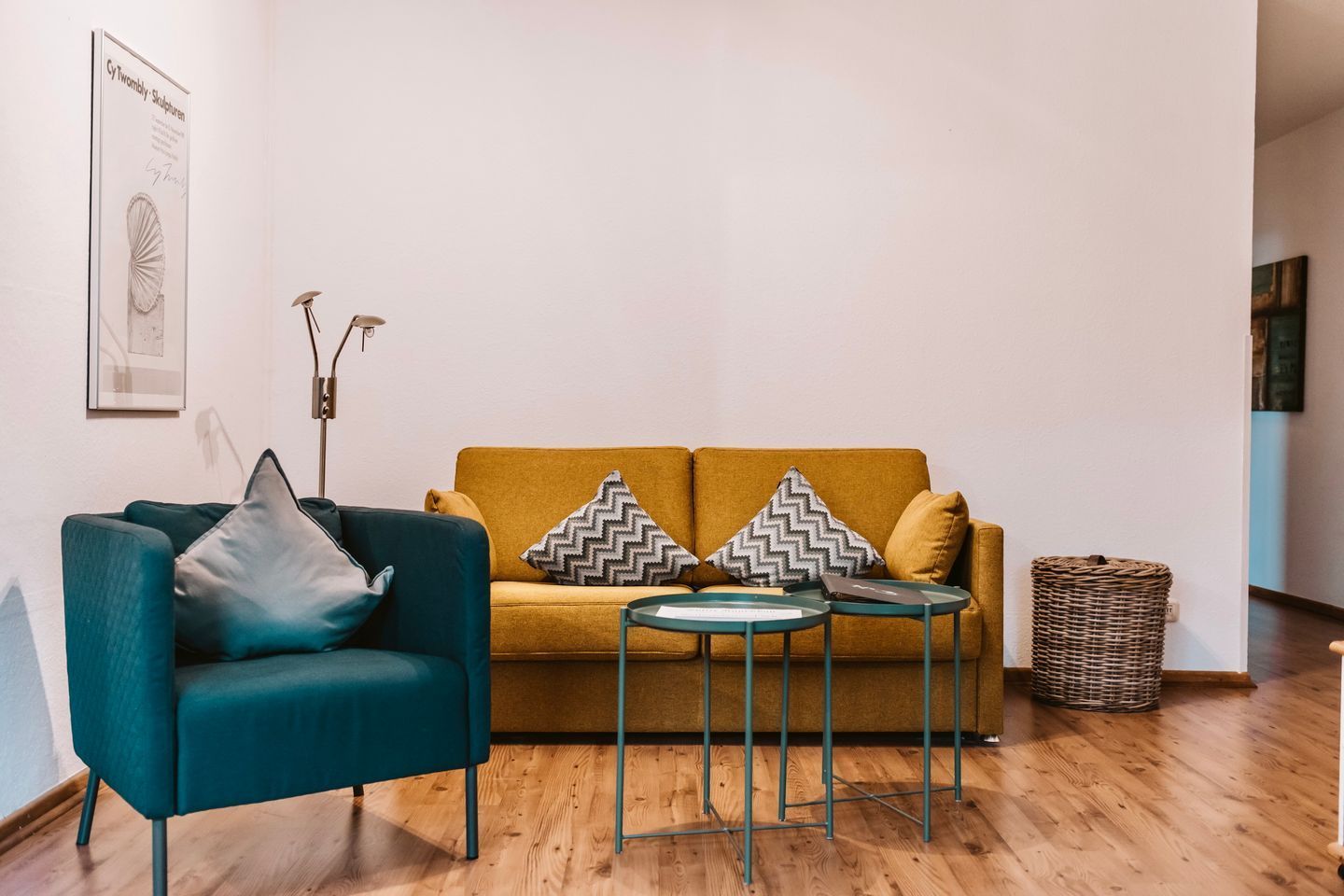 Een woonkamer met een smaragdgroene fauteuil, een mosterdgele bank, twee kleine ronde tafels en een rieten mand.