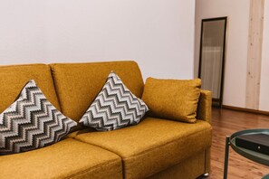 A mustard-colored sofa with patterned pillows and a glass coffee table in a room with wooden flooring and a door in the background. - Alter Konsumverein, App No. 3 - Cozy apartment in the Alter Konsumverein at (Sylt)