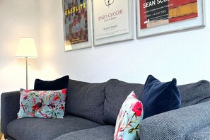Ein graues Sofa mit floralen und gemusterten Kissen, einer Stehlampe und gerahmten Bildern an der Wand.