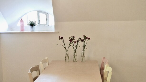 Et spisebord med fire glasvaser med blomster, et vindue med udsigt og en hylde med en lyserød flaske.