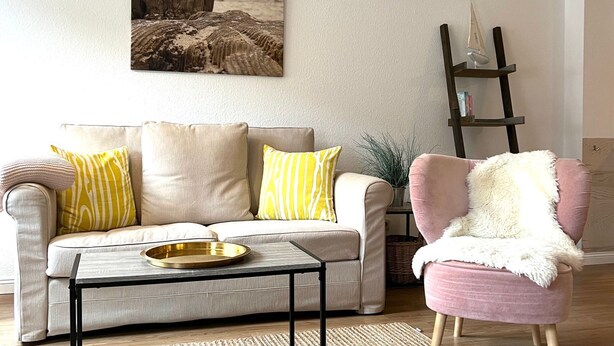 Een woonkamer met een beige bank, een salontafel, een roze fauteuil en een boekenplank.