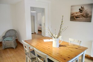 A dining area with a wooden table, white chairs, a wicker chair, and a vase with branches. - Alter Konsumverein, App No. 4 - Cozy Apartment at the Alter Konsumverein on (Sylt)