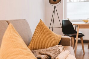 Un canapé avec un coussin texturé et une couverture, une lampe sur pied et un fauteuil dans une pièce dotée d’une fenêtre et d’une table.