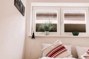 Ein Schlafzimmer mit einem Bett, Kissen, einem Fenster mit Jalousien, einer Topfpflanze und einer Wandlampe.