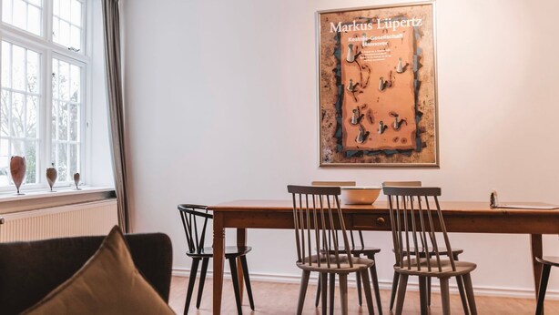 A dining area with a table and chairs, a window with curtains, and a framed artwork on the wall. - Alter Konsumverein, App No. 5 — An Oasis of Well-Being: Alter Konsumverein App No. 5 on Sylt (Sylt)