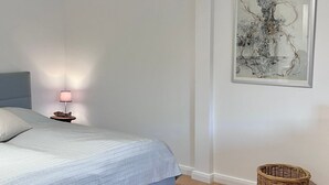 A bedroom with a bed, a nightstand with a lamp, a framed abstract artwork, and a wicker basket. - Alter Konsumverein, App No. 2 - Your Comfortable Apartment at Alter Konsumverein (Sylt)