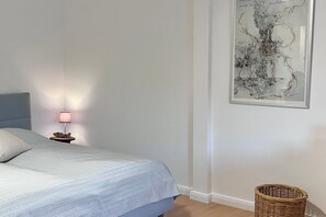 Ein Schlafzimmer mit einem Bett, einem Nachttisch mit Lampe, einem gerahmten abstrakten Kunstwerk und einem Korbwägelchen.
