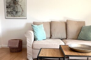 Een woonkamer met een beige bank, een klein houten bijzettafeltje, een rieten mand en een abstract schilderij in een lijst aan de muur.