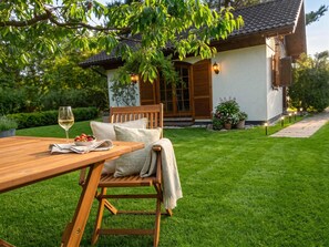 A wooden outdoor dining table with a glass of wine and a bowl of fruit, a wooden bench with cushions, and a house with a tiled roof and a garden.