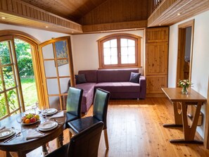 Ein Zimmer mit Holzboden, einem violetten Sofa, einem Esstisch mit Stühlen und einem Fenster mit Blick auf Grünflächen.