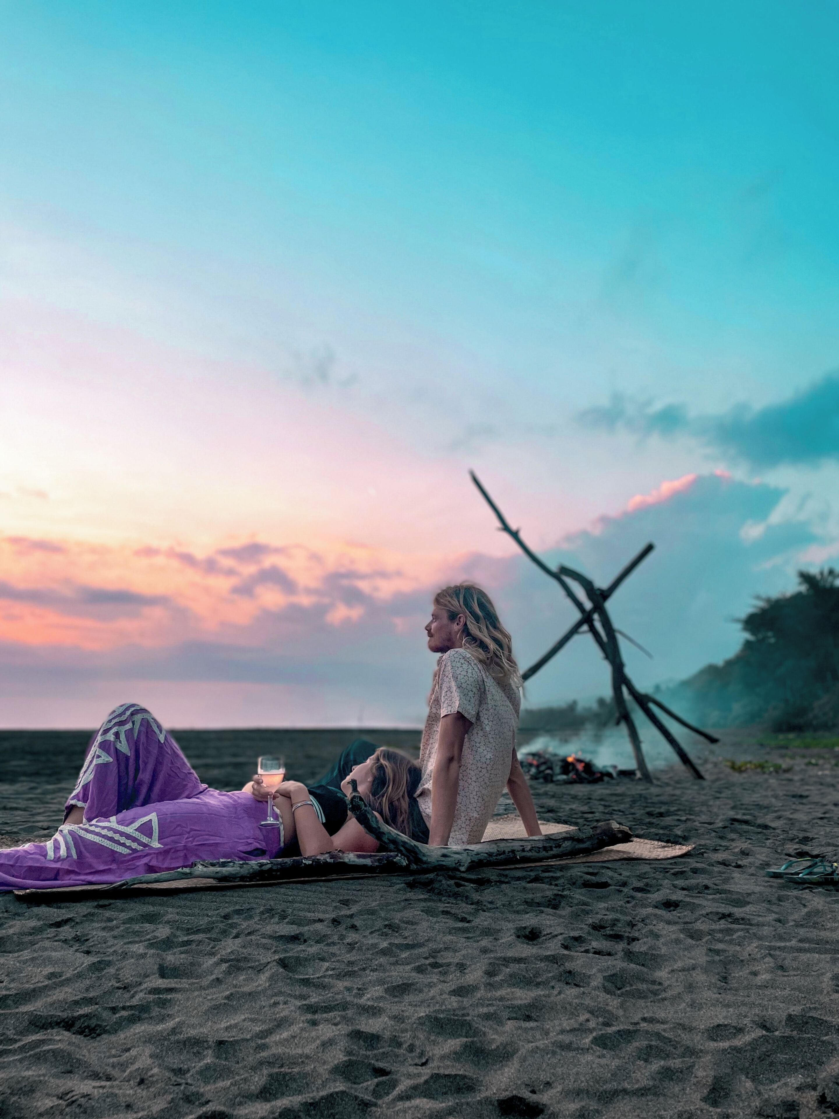 Eine Strandansicht mit zwei Personen, eine liegt auf einer Matte, die andere steht, mit einer Holzkonstruktion im Hintergrund.