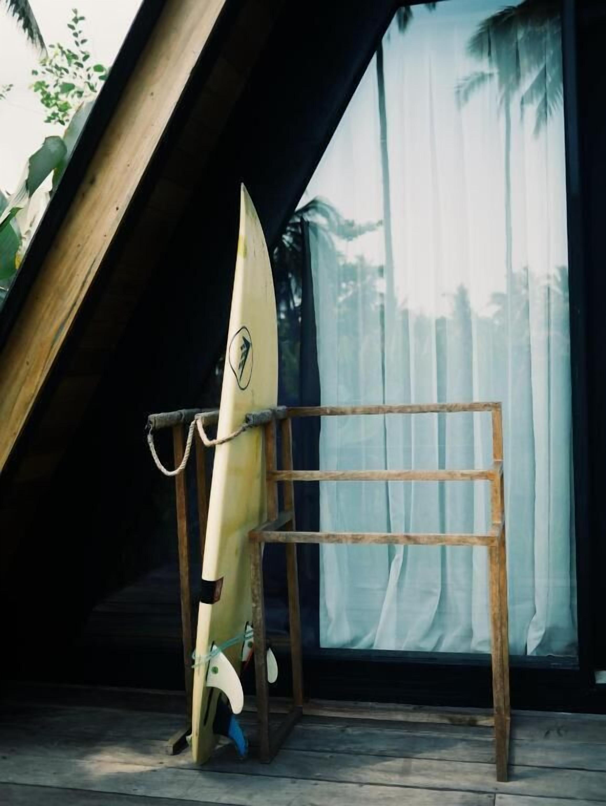 A surfboard leaning against a wooden ladder in a room with a large window and curtains.