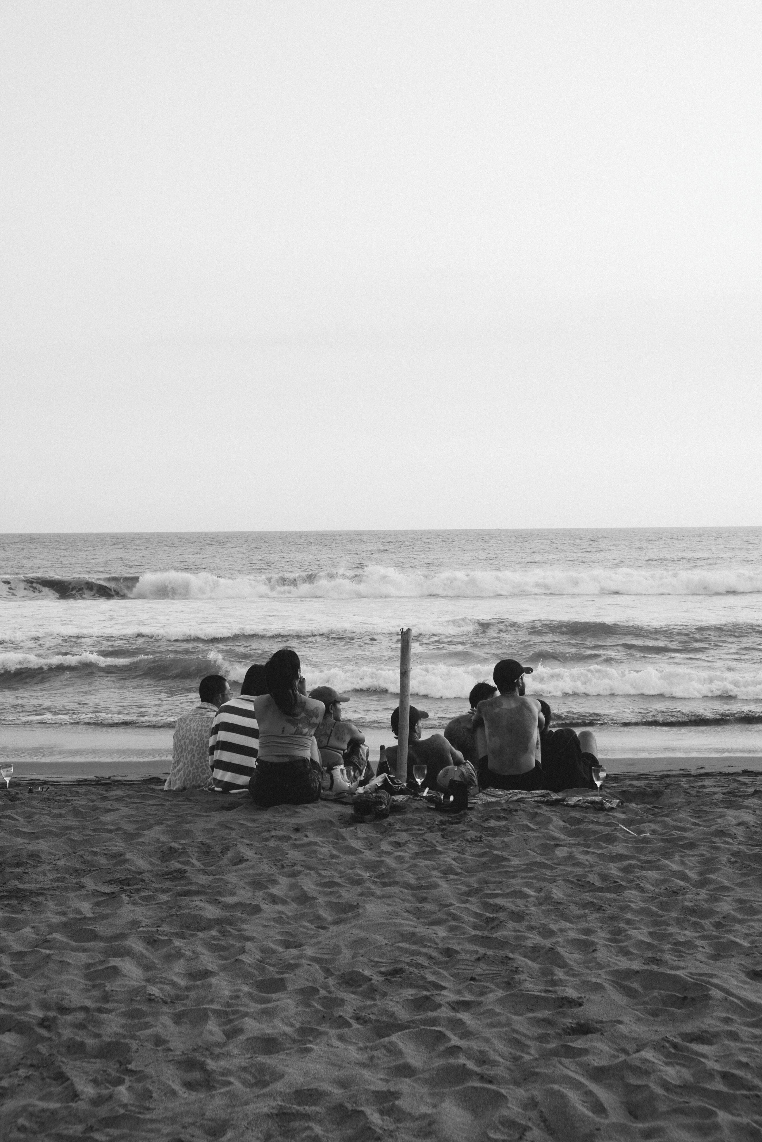 A beach scene with people sitting on the sand, some with drinks and bags, facing the ocean.
