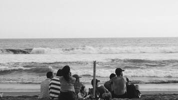 A beach scene with people sitting on the sand, some with drinks and bags, facing the ocean.
