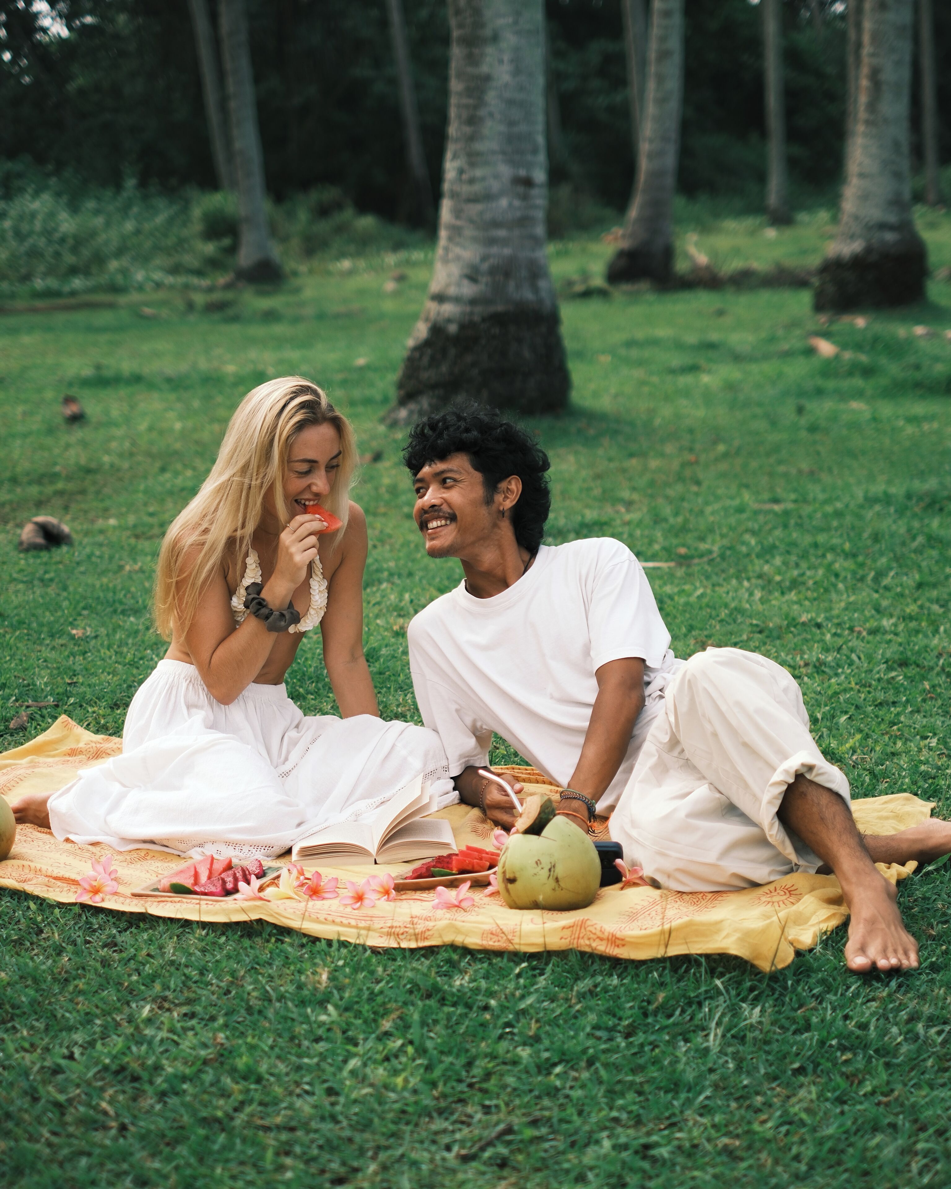 Two people sitting on a blanket outdoors with a book, a coconut, and some food items.