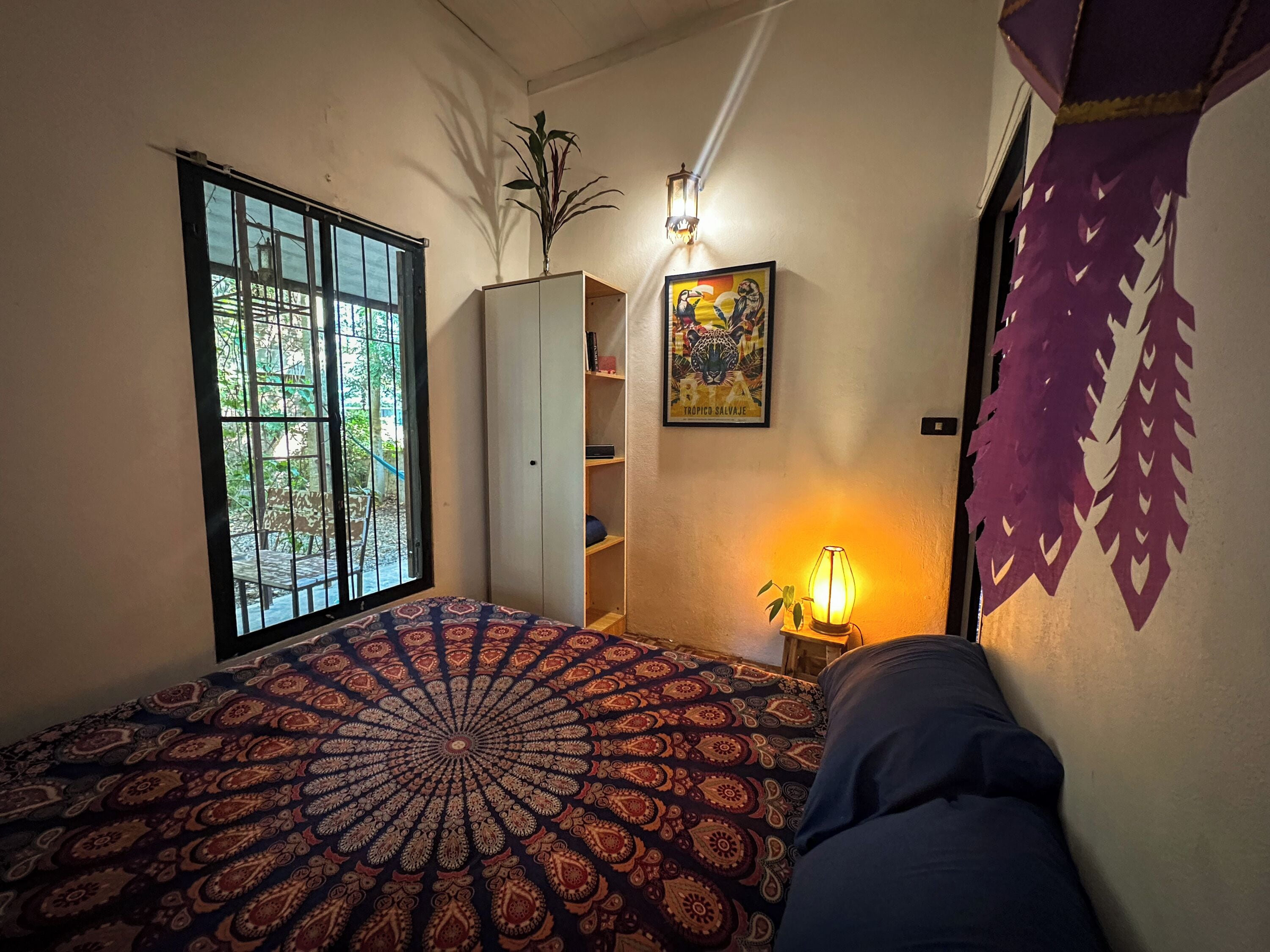 A room with a large window, a bed with a patterned bedspread, a wall-mounted shelf, a wall lamp, and a framed picture.