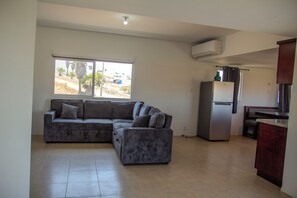 A living room with a sectional sofa, a refrigerator, and a window with a view of a residential area. - Casa Vista del Mar  Relax, Unwind & Watch the Sunset (Playas de Rosarito)