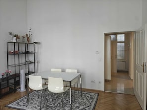 A room with a white table, four white chairs, a patterned rug, a metal shelving unit with various items, and a door leading to another room. - Elegant Altbau near Praterstern
Direct Subway to Center (Wien)
