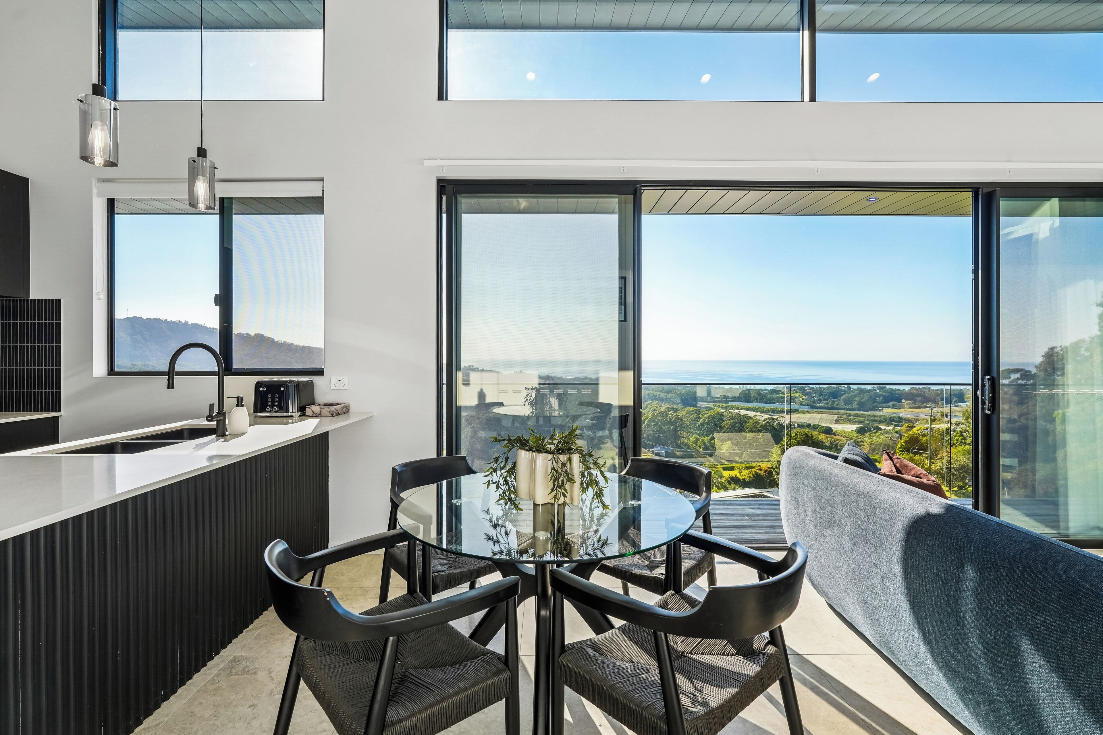A modern kitchen with a large island, a dining area with a glass table and chairs, and a balcony with a view of the landscape.